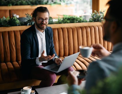 Young happy entrepreneur using laptop while talking to coworker during business meeting in cafe. Happy businessman working on laptop while talking to colleague during a meeting in cafe.